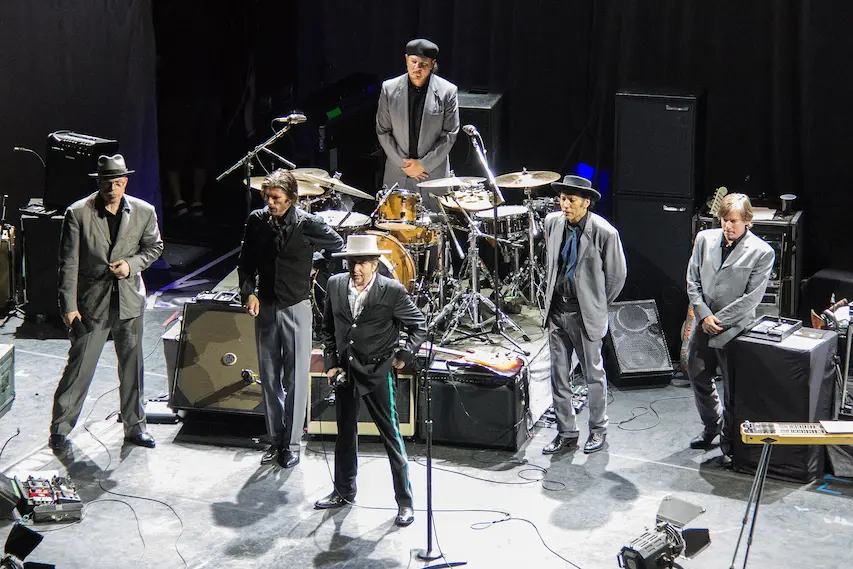 Bob Dylan et son groupe saluant le public en fin de concert sur scène, habillés en costumes vintage.