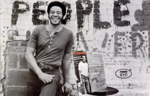 Bill Withers souriant devant un mur tagué, pochette de l’album Just As I Am visible au premier plan, ambiance vintage