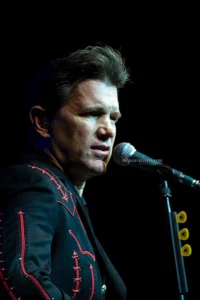 Chris Isaak en concert, gros plan au micro, vêtu d'une veste noire brodée de rouge.