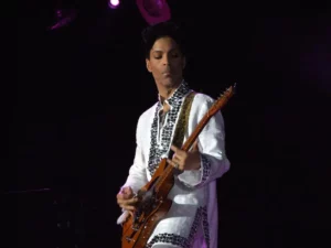 Prince sur scène en costume blanc, jouant un solo sur une guitare orange custom, éclairage de concert