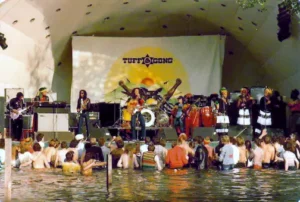 Bob Marley et les Wailers en concert devant un public debout dans l’eau, scène avec percussions et choristes