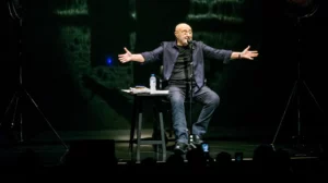 Phil Collins assis sur un tabouret, chantant avec intensité et les bras écartés pendant un concert