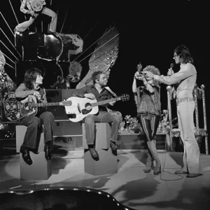 Le groupe Stealers Wheel jouant des guitares sur scène avec un décor élaboré, un homme debout tenant un micro, et une femme à côté.