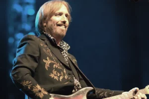 Tom Petty souriant sur scène, jouant de la guitare avec une veste brodée et une chemise à motifs