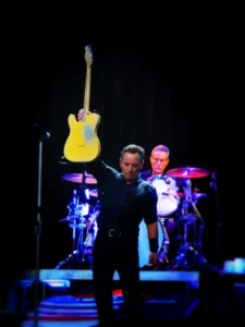 Bruce Springsteen lève sa guitare jaune en concert, éclairé sur fond noir avec son batteur en arrière-plan