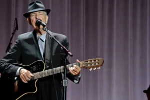 Leonard Cohen chantant sur scène, les yeux fermés, en costume noir et chapeau, avec une guitare acoustique