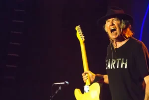 Neil Young en pleine performance scénique avec une guitare électrique jaune et un t-shirt noir marqué "Earth"