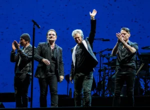 Les membres de U2 sur scène, saluant le public en concert sous des lumières bleues
