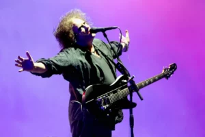 Robert Smith, chanteur et guitariste de The Cure, interprétant Boys Don’t Cry sur scène, bras ouverts, sous un éclairage violet et rose.