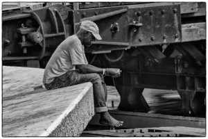 Homme assis pieds nus sur un quai, regardant l’eau, en noir et blanc