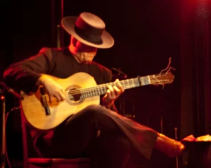Guitariste flamenco vêtu de noir avec un chapeau traditionnel, jouant sur une guitare classique sous un éclairage rouge, dans l’esprit des Gipsy Kings.