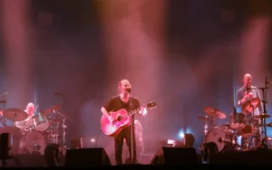Thom Yorke chantant et jouant de la guitare acoustique sur scène avec Radiohead, accompagné des batteurs, sous des lumières rouges et bleutées.